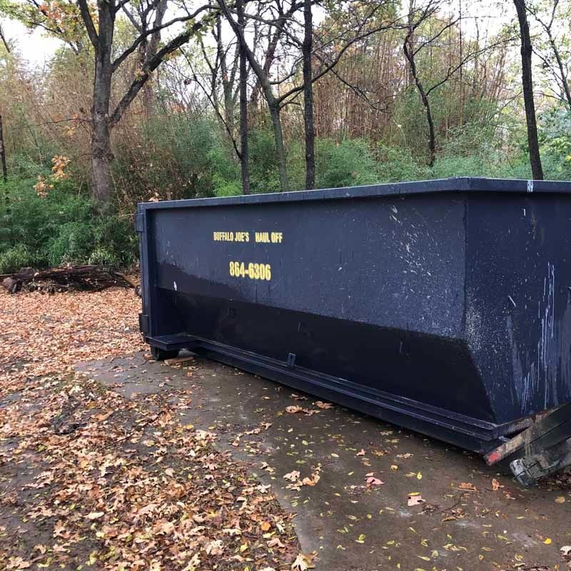 Large dark blue dumpster in a wooded area covered with fallen leaves and wet ground.