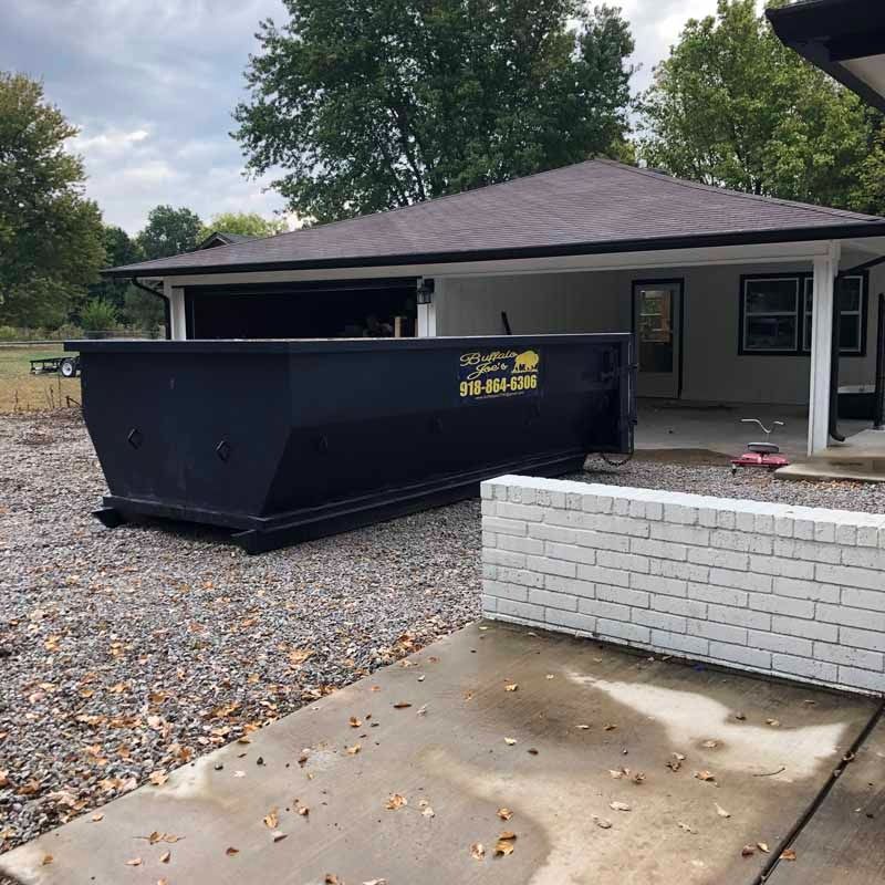 Large black dumpster beside a house and white brick wall on a gravel driveway, with fallen leaves scattered around.