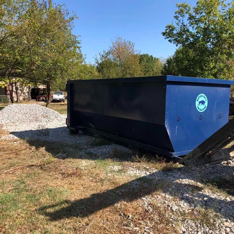 Blue dumpster on gravel beside trees under a clear sky.