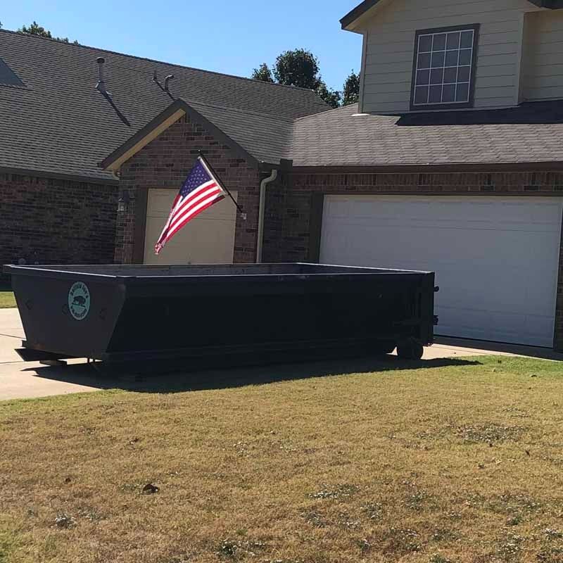 Large black dumpster on a suburban lawn in front of a house with an American flag.