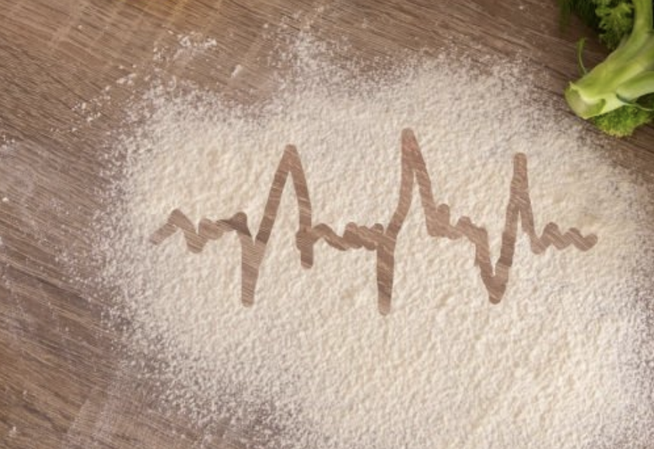 A wooden surface covered in a light dusting of white flour, with a heart rate (EKG) pulse line traced clearly through the center. A small piece of fresh green broccoli is visible in the top right corner.
