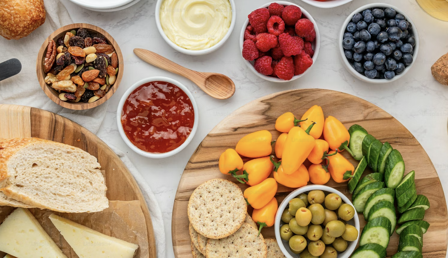 A top-down view of a marble table covered in various healthy snacks, including small bowls of raspberries, blueberries, olives, and jam, alongside sliced bread, cheese wedges, crackers, small orange peppers, and sliced cucumbers.