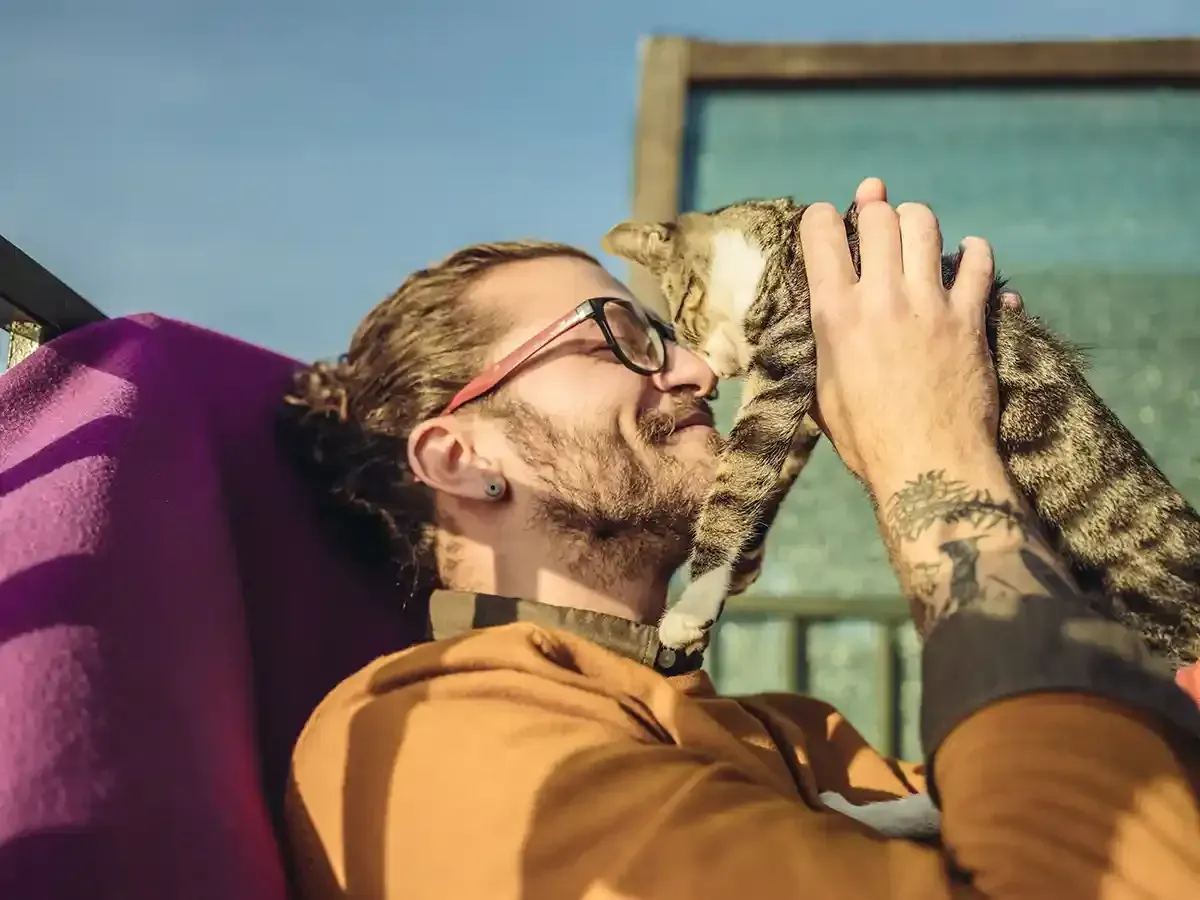 Person in glasses kisses a tabby cat outdoors on a sunny day