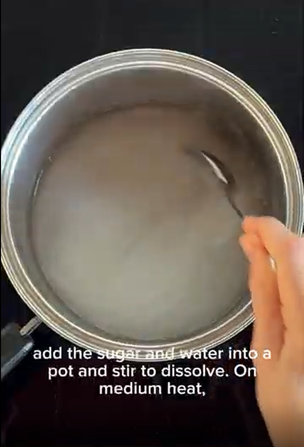 A hand stirs sugar and water in a metal pot on a stovetop. Text overlay instructs to heat on medium.