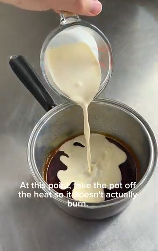 Cream being poured from a glass measuring cup into a saucepan containing a dark liquid on a metal counter.