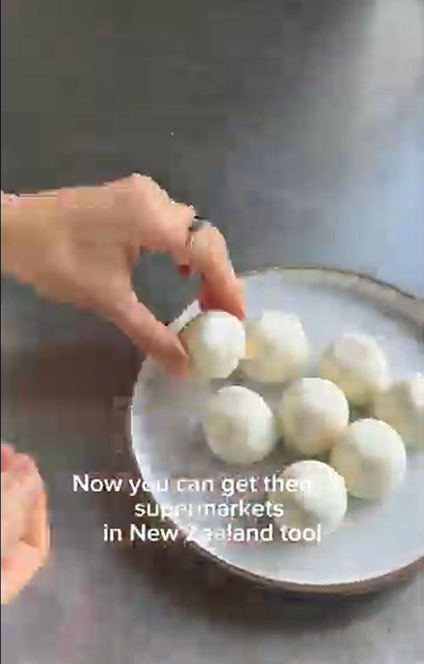 A hand places a white, ball-shaped food item onto a plate with several others, captioned about availability in NZ.