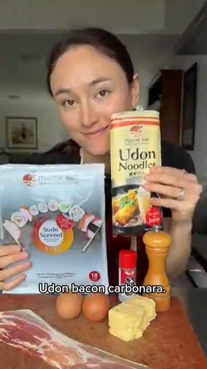 A person holding packs of Udon noodles and sushi seaweed, with eggs, bacon, butter, and seasonings on a counter.