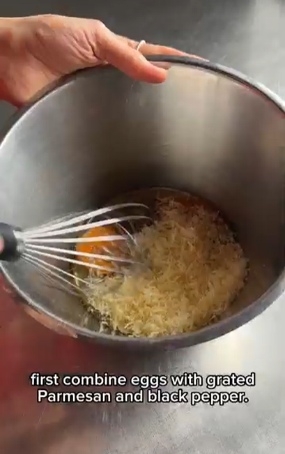 A hand uses a whisk to combine eggs, grated Parmesan, and black pepper in a stainless steel mixing bowl.