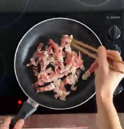 Hands using wooden chopsticks to stir raw bacon strips in a black frying pan on a stovetop.