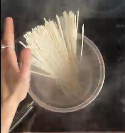 A hand placing a bundle of dry white noodles into a pot of boiling water on a stovetop.