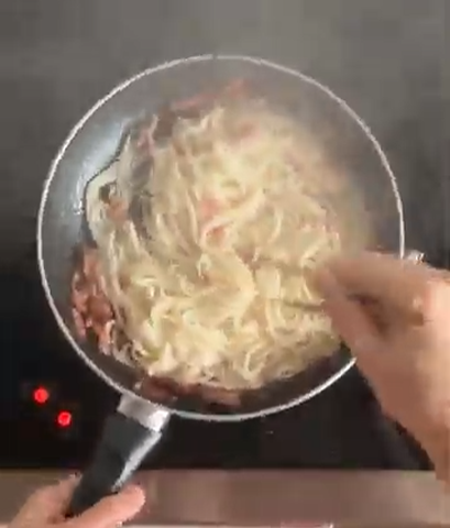 A hand uses tongs to stir pasta with bacon and a creamy sauce in a frying pan on a stovetop.