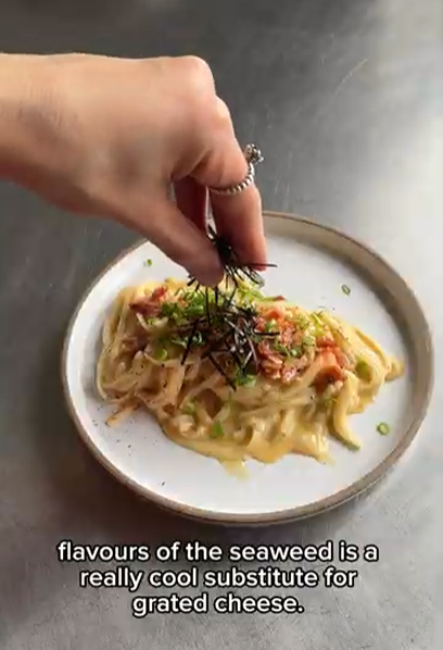 A hand sprinkles shredded seaweed onto a plate of creamy pasta. Text overlay mentions seaweed as a cheese substitute.