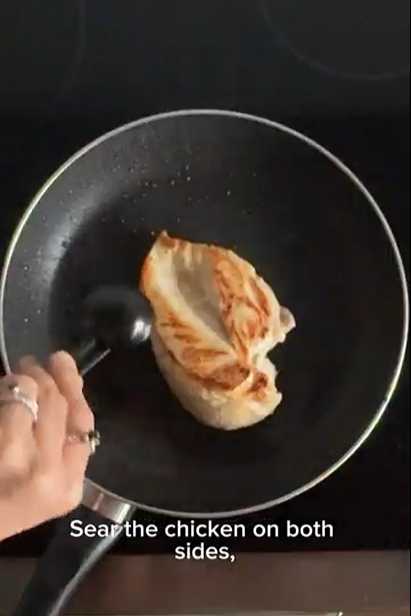 A hand uses black tongs to sear a piece of chicken in a frying pan on a stovetop.