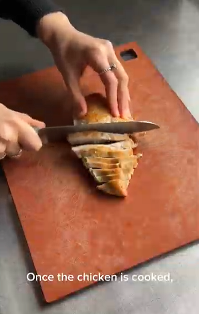 A person's hands use a knife to slice a cooked chicken breast on a brown cutting board.
