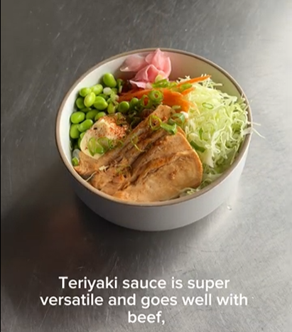 A bowl of food with teriyaki-glazed meat, edamame, shredded cabbage, and pickled ginger on a metal surface.