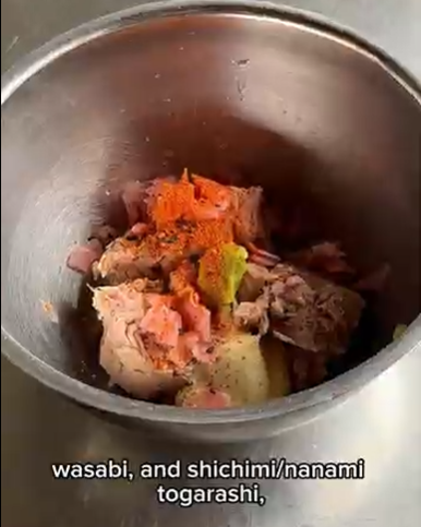 A metal bowl containing tuna, diced red onion, a dollop of wasabi, and a dusting of red shichimi togarashi seasoning.