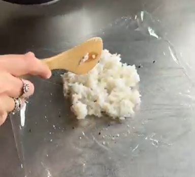 A hand uses a wooden spatula to press and spread a pile of cooked white rice onto a sheet of clear plastic wrap.