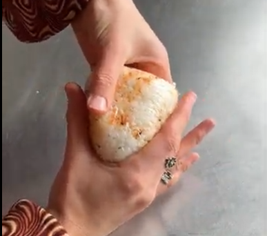 Hands gently shaping a triangular rice ball, likely onigiri, on a smooth surface.