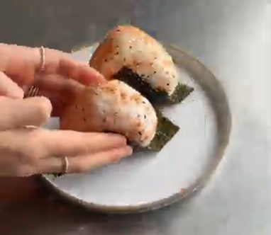 Hands place a grilled, triangular onigiri topped with sesame seeds and nori onto a white plate holding another one.
