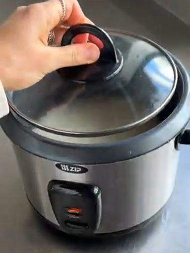 Hand holding lid on a stainless steel electric rice cooker with black trim