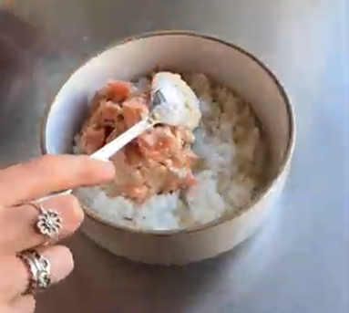 A hand with rings scoops pink tuna onto white rice in a bowl on a metal surface