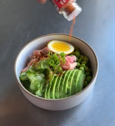 A bowl of salad with avocado, cucumber, peas, sliced egg, and greens, topped with dressing being poured in.