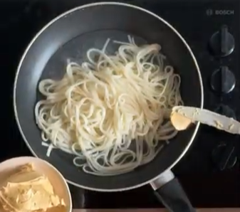 Overhead view of a pan with noodles on a stovetop, with a spatula and bowl of ingredients nearby