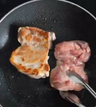 Two pieces of meat sizzling in a black frying pan, one browned and one raw being turned with tongs