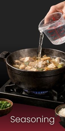 A hand pours water from a measuring cup into a black pot of cooking stew on a gas stove. 