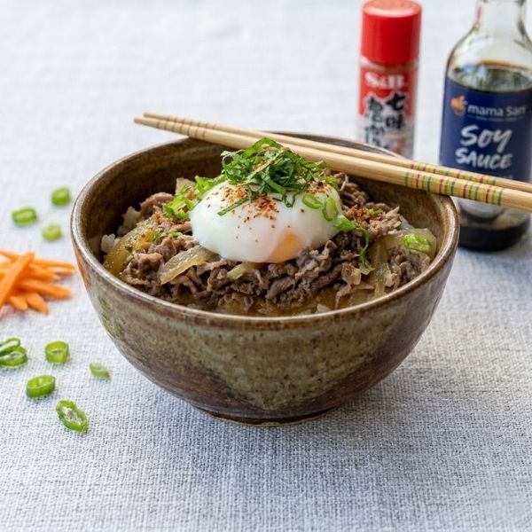 Bowl of beef rice topped with a poached egg, green onions, and chopsticks, with soy sauce behind it