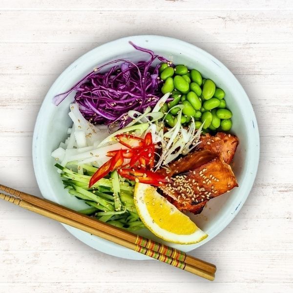 Bowl of colorful poke with salmon, edamame, cabbage, cucumber, radish, and chopsticks on a light table