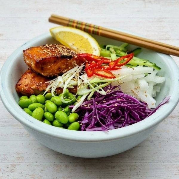 Rice bowl with salmon, edamame, cabbage, cucumber, and lemon, topped with sliced red peppers and chopsticks
