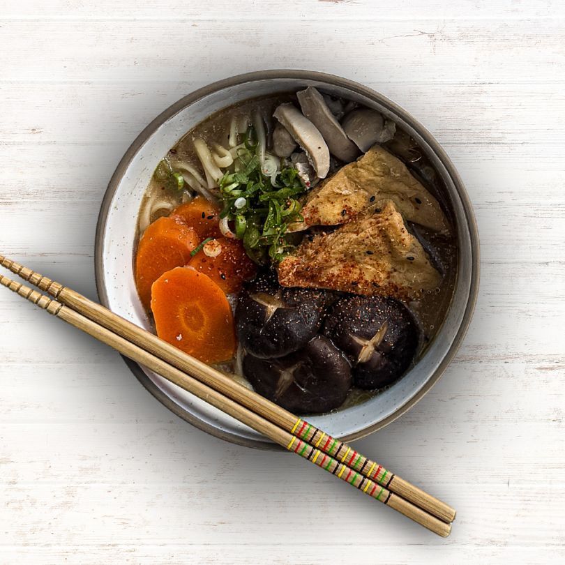 Bowl of vegetable noodle soup with chopsticks on a light wooden table.