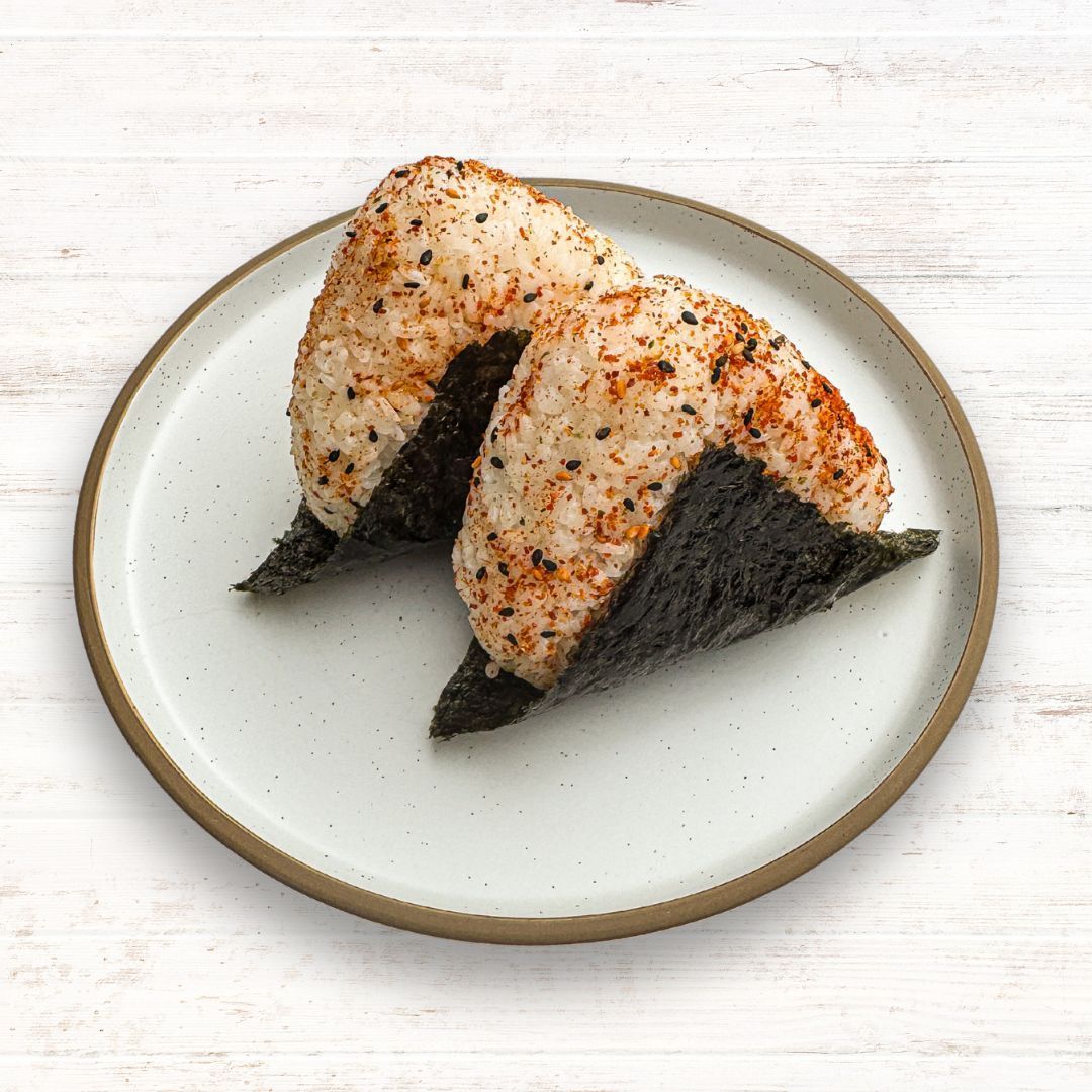 Two triangular tuna mayo rice balls topped with seasoning and wrapped in nori, served on a speckled plate.