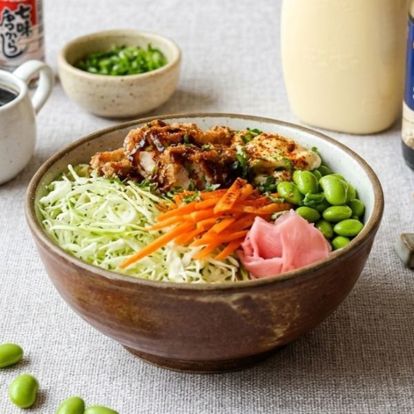 Japanese-style poke bowl with shredded cabbage, carrots, edamame, pickled ginger, and glazed chicken on a table