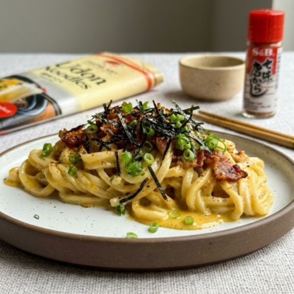 Plated creamy pasta topped with herbs and seaweed, with a cookbook and seasoning bottle in the background