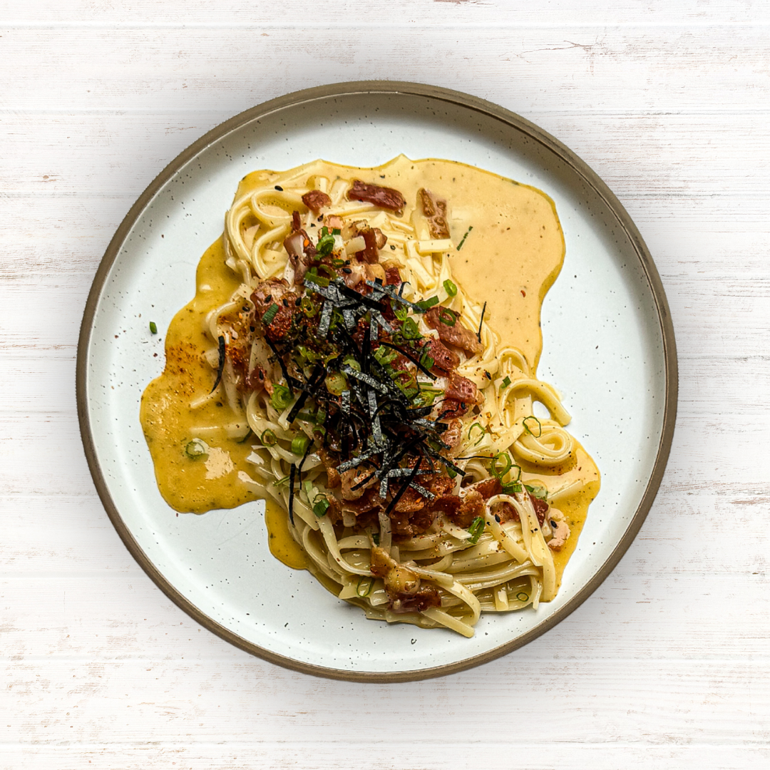 A serving of creamy, saucy pasta topped with crispy bits, chopped green onions, and thin strips of dark seaweed on a plate.