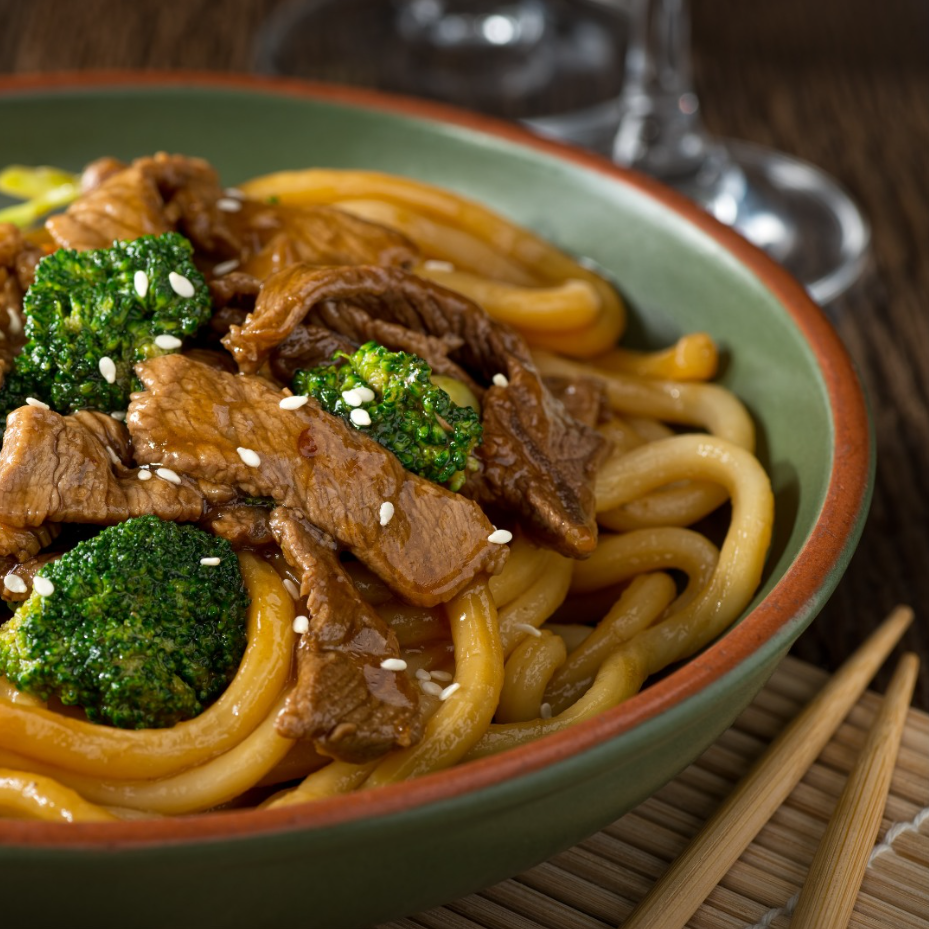 Beef and broccoli udon noodles in a green bowl, with chopsticks and a wooden placemat.