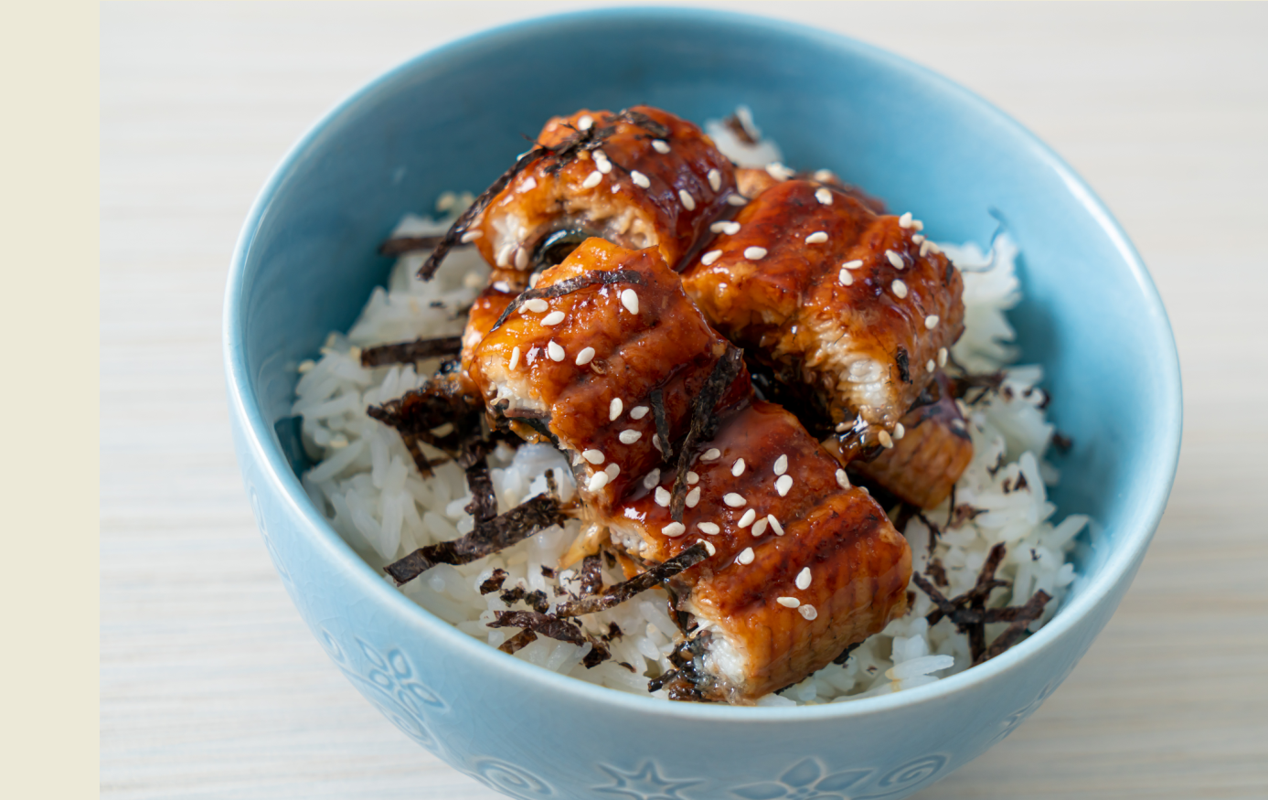 Bowl of white rice topped with glazed eel, sesame seeds, and seaweed in a light blue bowl.
