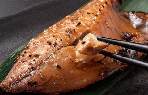 A cooked, glazed fish fillet resting on a green leaf, with a piece being lifted by black chopsticks.