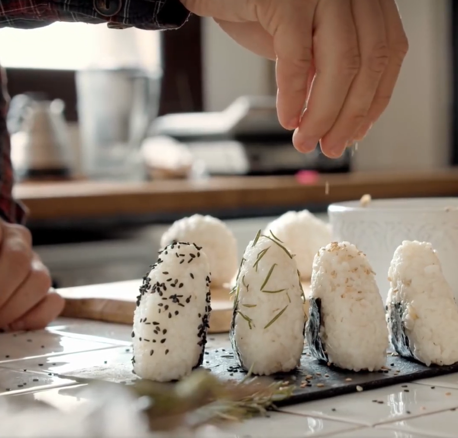 Person sprinkling seeds on rice balls, kitchen setting.