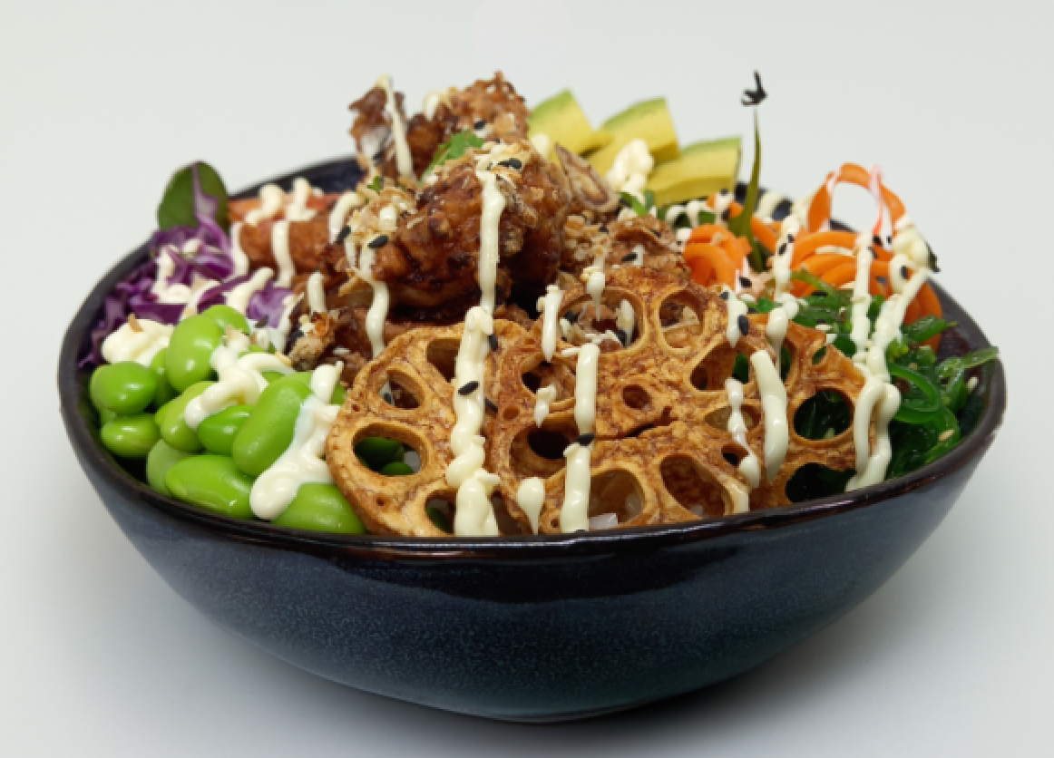 A colorful poke bowl with tempura, avocado, and vegetables, drizzled with white sauce, in a dark bowl.