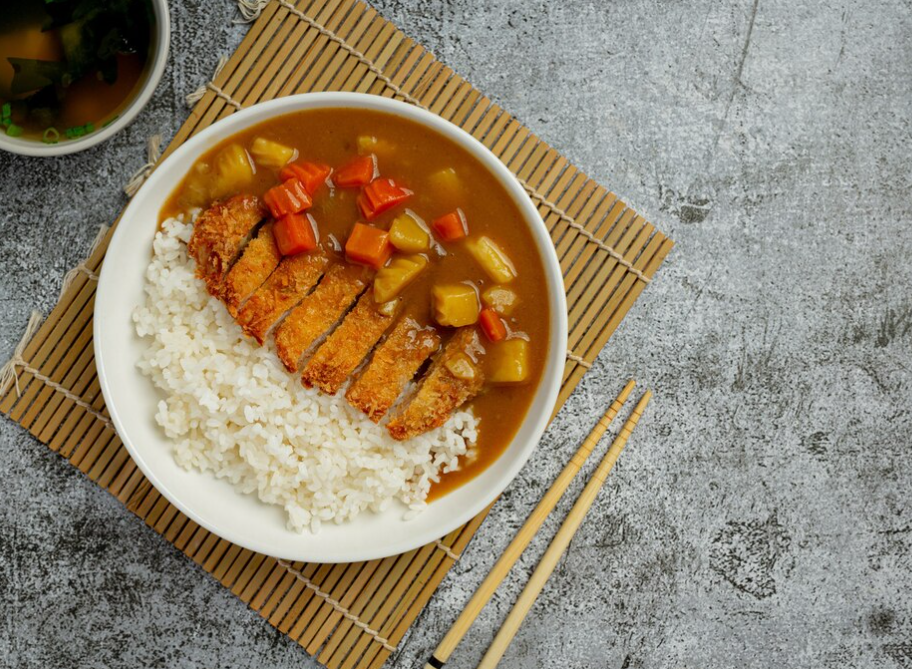 Japanese curry with tonkatsu, rice, and vegetables in a white bowl, with chopsticks and soup on a bamboo mat.