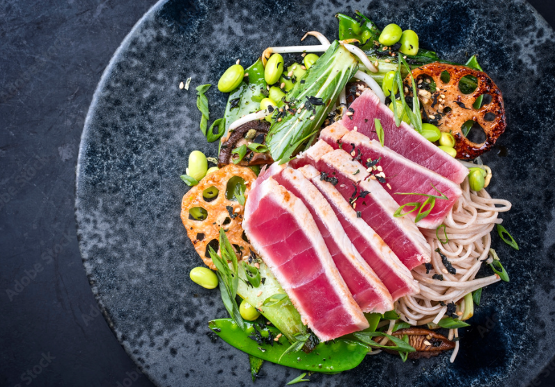 Seared tuna over soba noodles with edamame and vegetables on a dark plate.