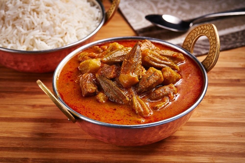Okra curry in a copper bowl next to a bowl of white rice, on a wooden table.