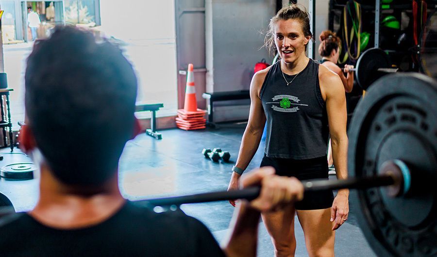 Gym coach standing in front of a barbell at a gym in Winter Park, FL, smiling while a lifter squats in the foreground.