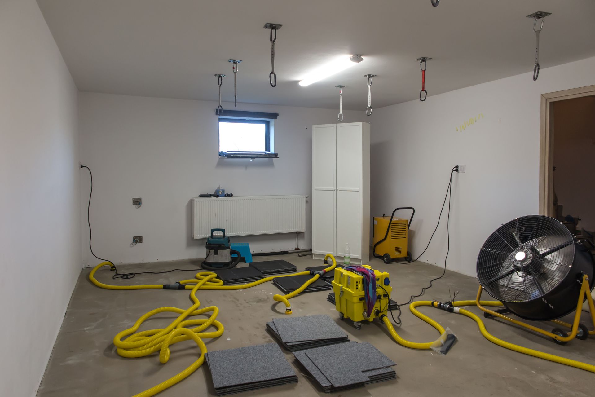 A room with dehumidifying equipment and water-damaged floor tiles. Yellow hoses, white walls, and a large fan.
