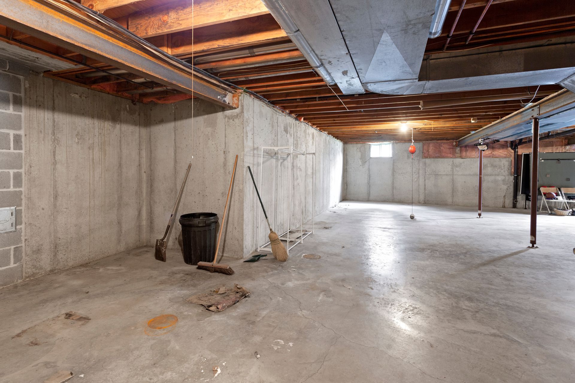 Basement with concrete walls and floor; exposed ceiling beams and ductwork; brooms and trash can.