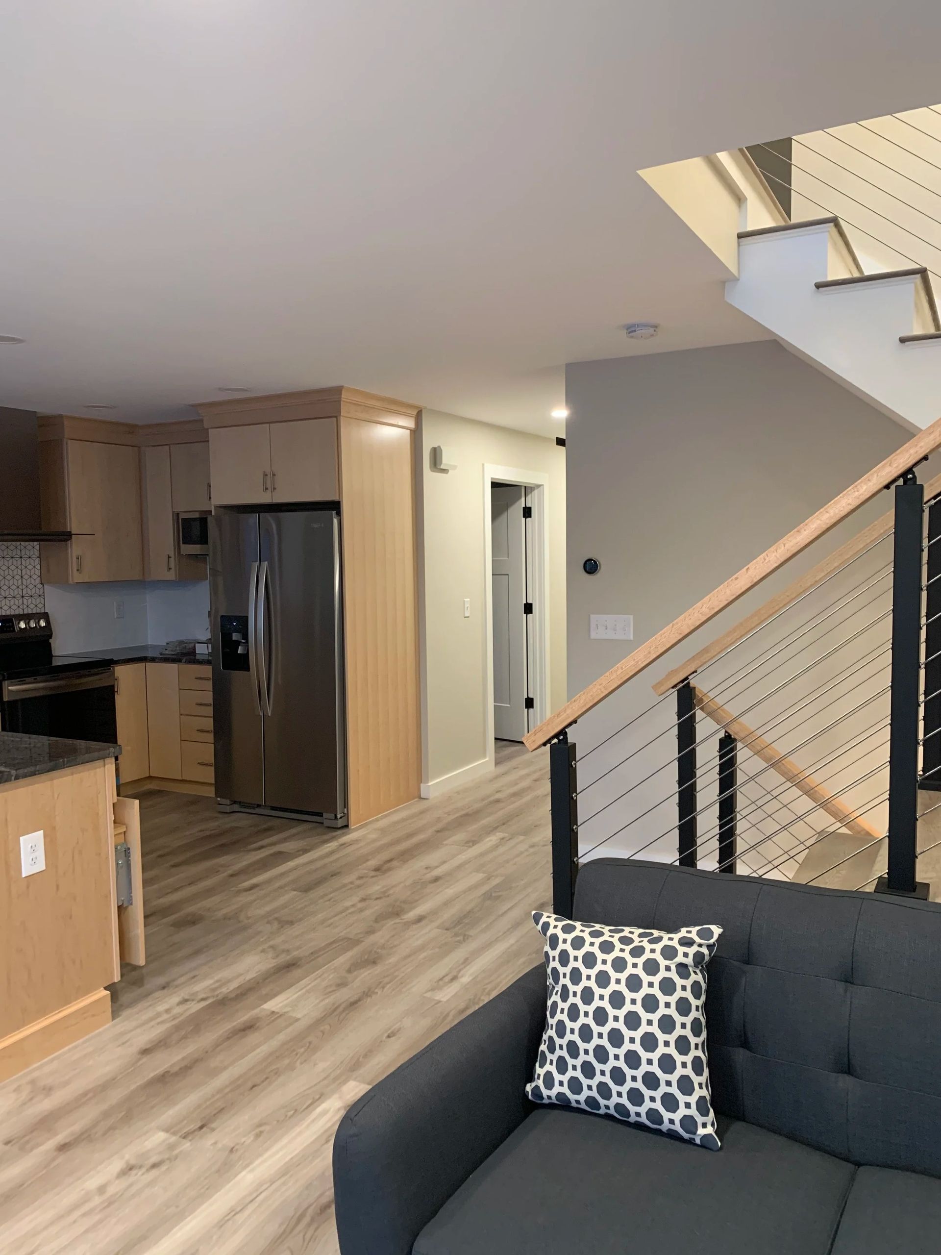 Modern kitchen and living room with stainless steel fridge, light wood cabinets, gray couch, and staircase.