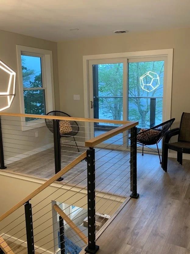 Modern stairwell with glass and cable railing, wood handrail, and black posts. Two chairs face a window.